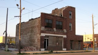 Roofless boarded up brick building, with piles of bricks fallen around it